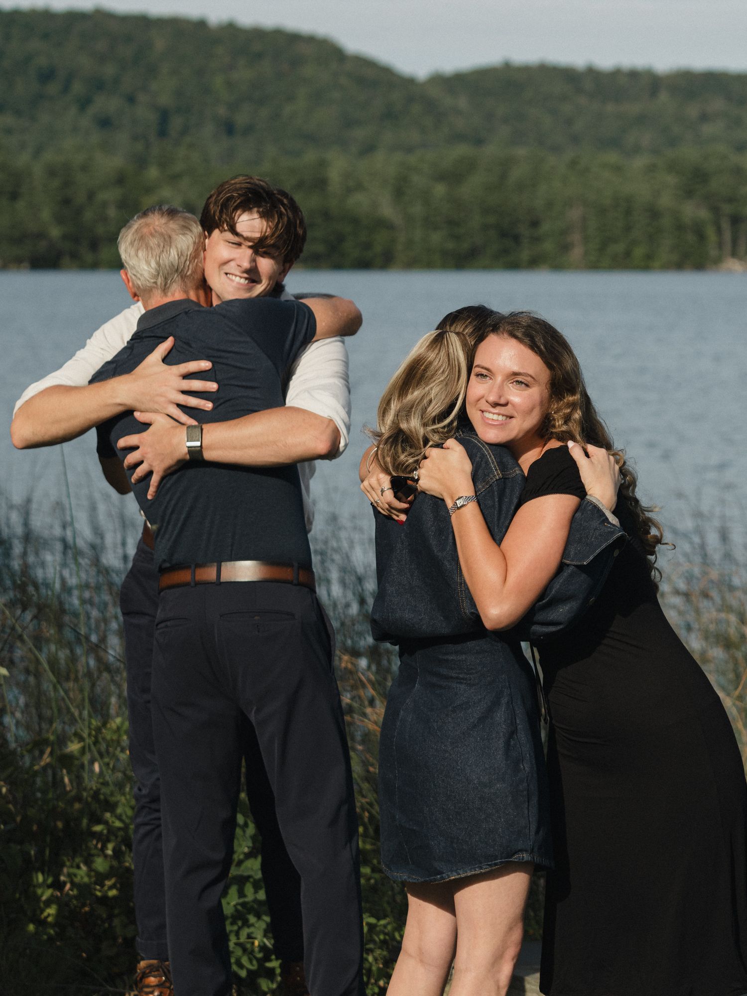 People sharing warm embraces outdoors against a scenic lake backdrop.