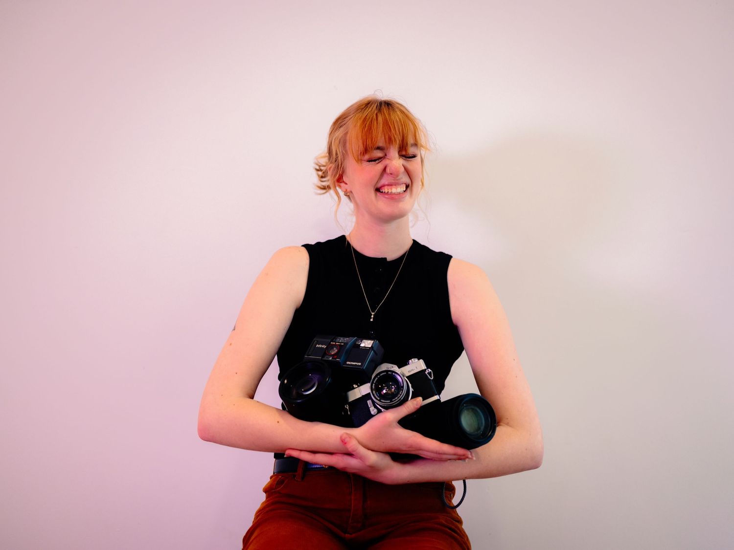 A photographer in a black sleeveless top and burgundy pants holds a vintage camera against a pink wall.
