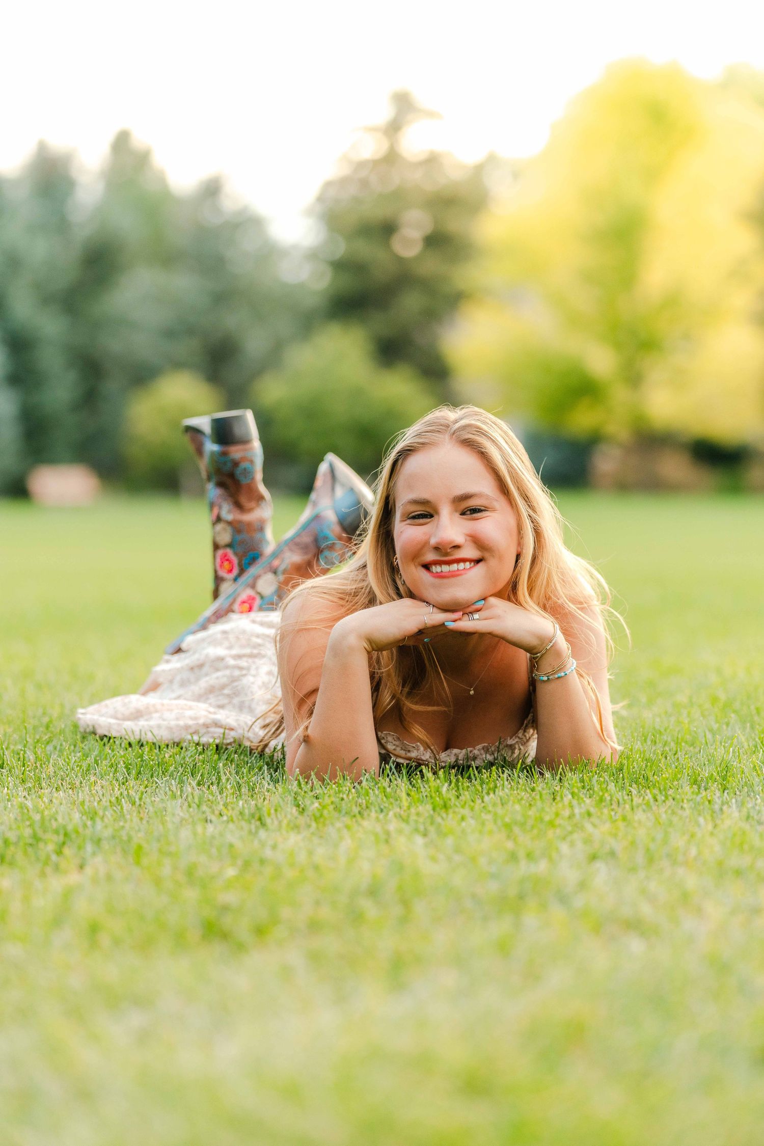 Smiling blonde woman lying on green grass in a sunny park wearing a floral dress and boots.
