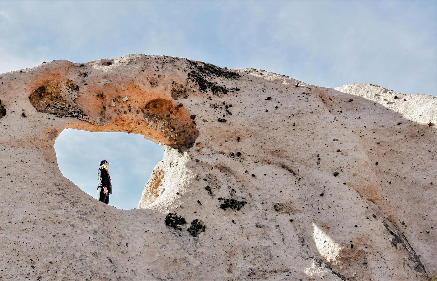 Person standing in a natural rock arch formation against a blue sky in an arid landscape.
