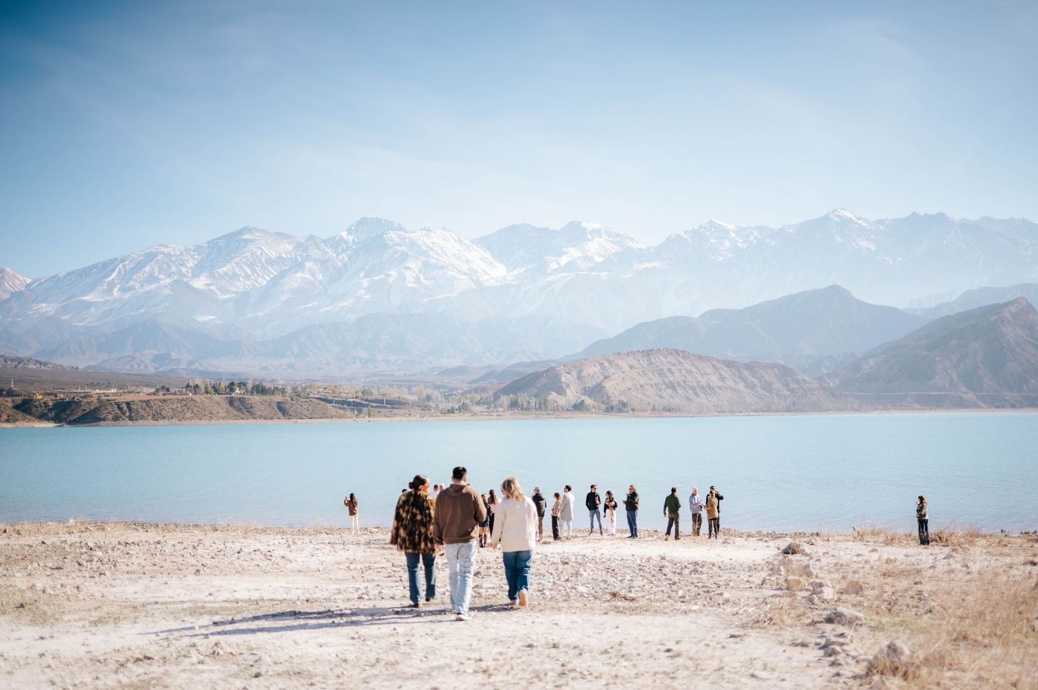 Wedding guests walking along a scenic lakeside path with snow-capped mountains in the background.