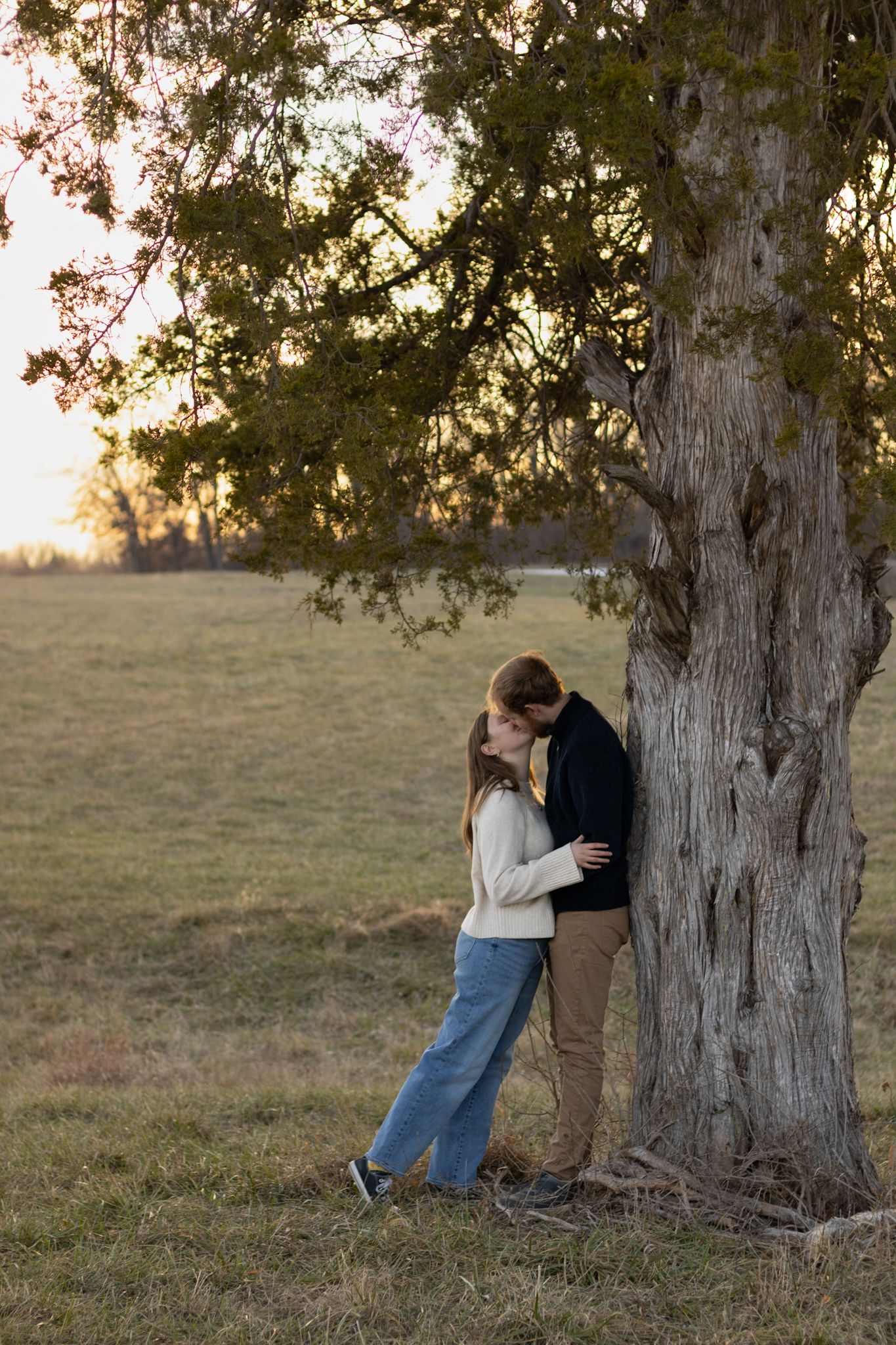 Couple kissing under a large tree in an open field at sunset, woman in white sweater and jeans, man in dark jacket.