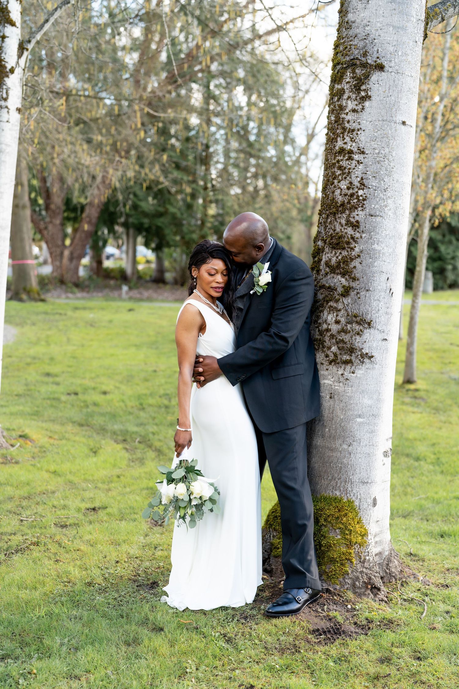 A bride and groom share a tender moment leaning against a tree in a lush green park on their wedding day.