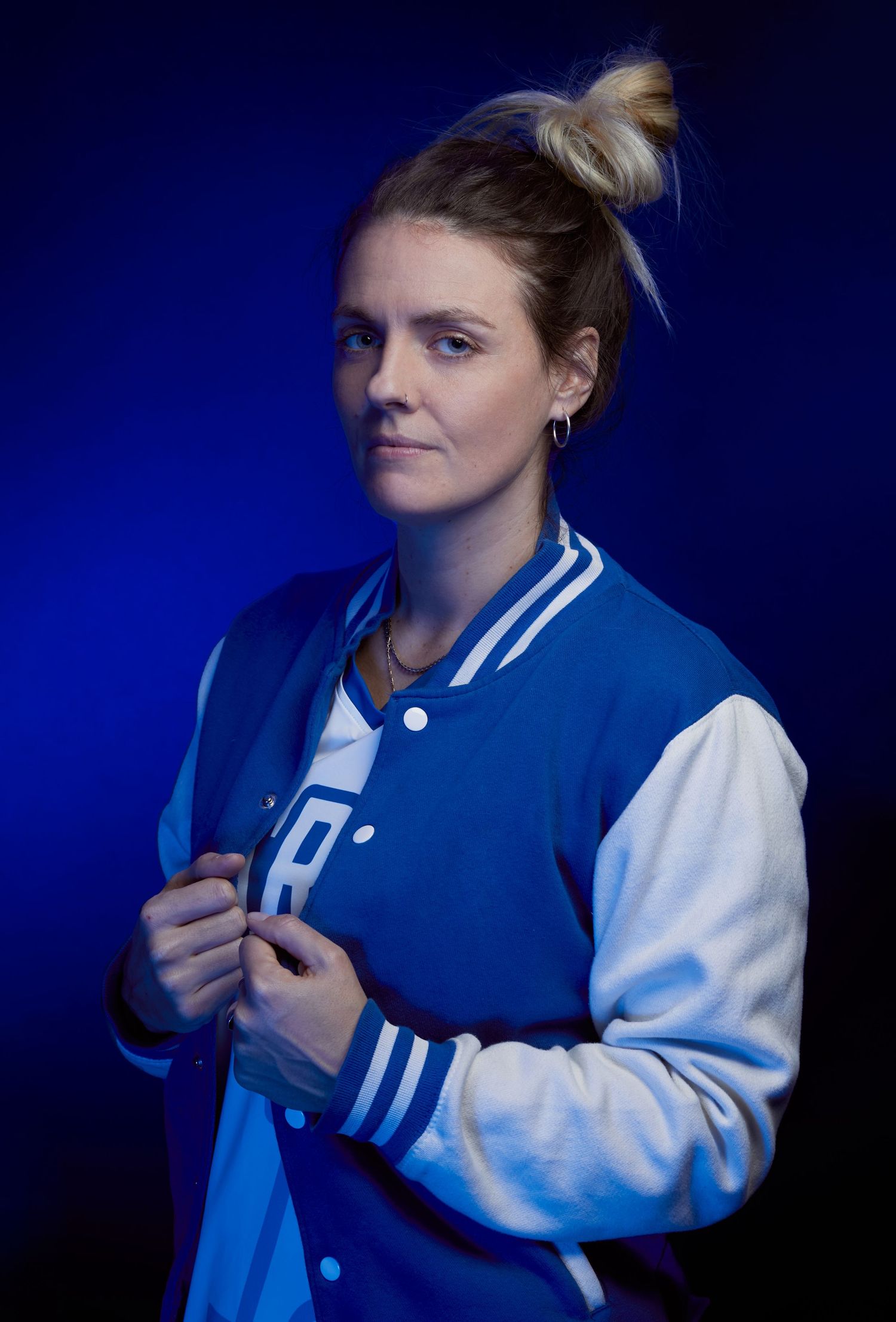 Woman in blue and white varsity jacket poses against dark blue background with hair in messy bun.