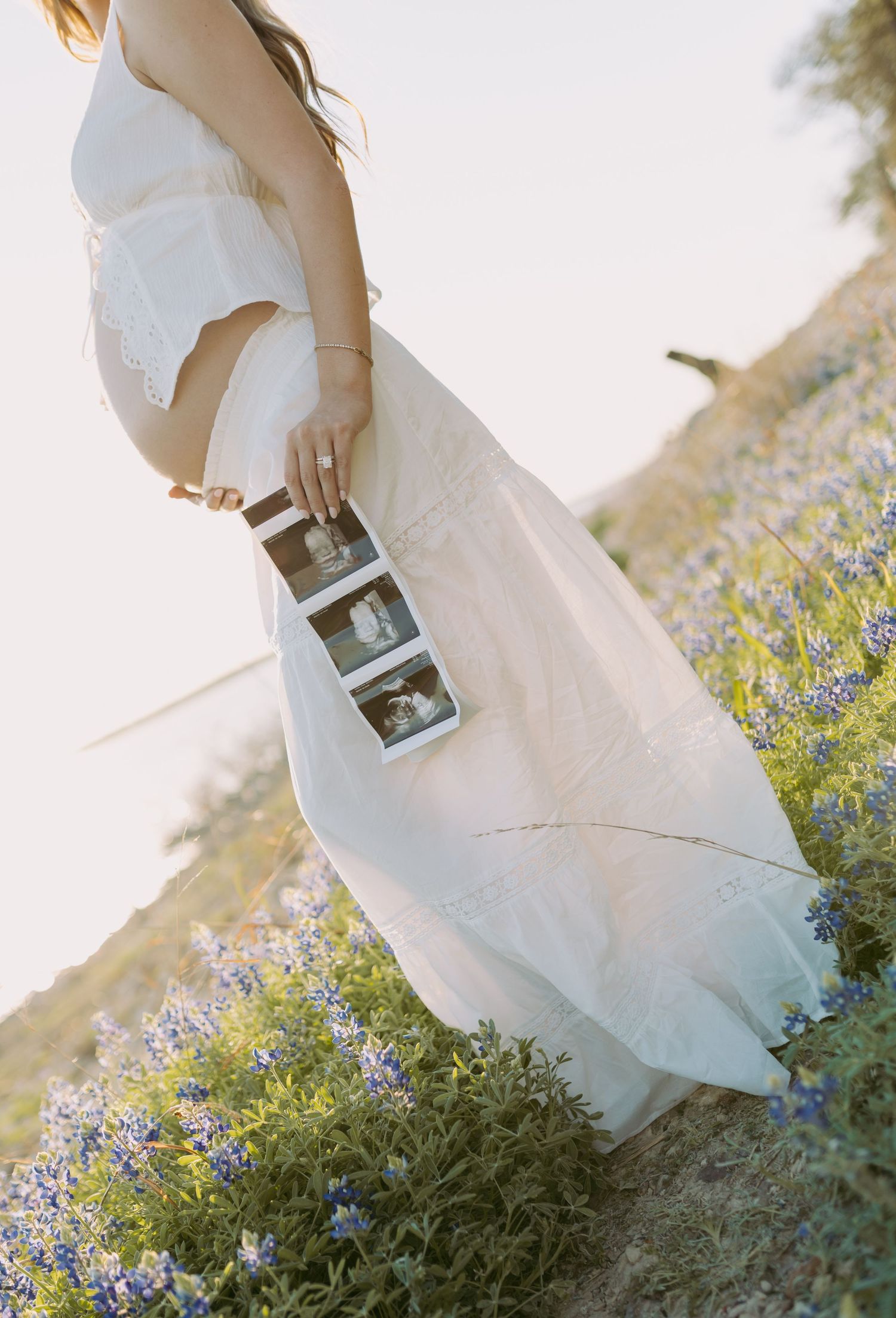 A person in a flowing white dress holds ultrasound photos while standing among blooming blue wildflowers at sunset.