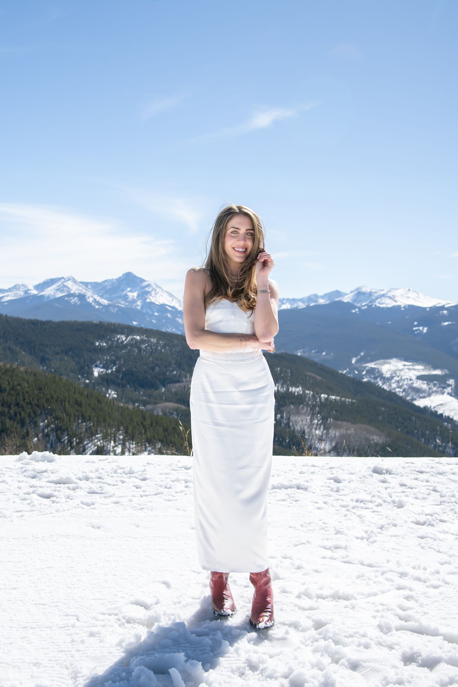 Fashionable cute woman in red boots and white dress standing on the snow with mountain peaks in the background in Vail Colorado.