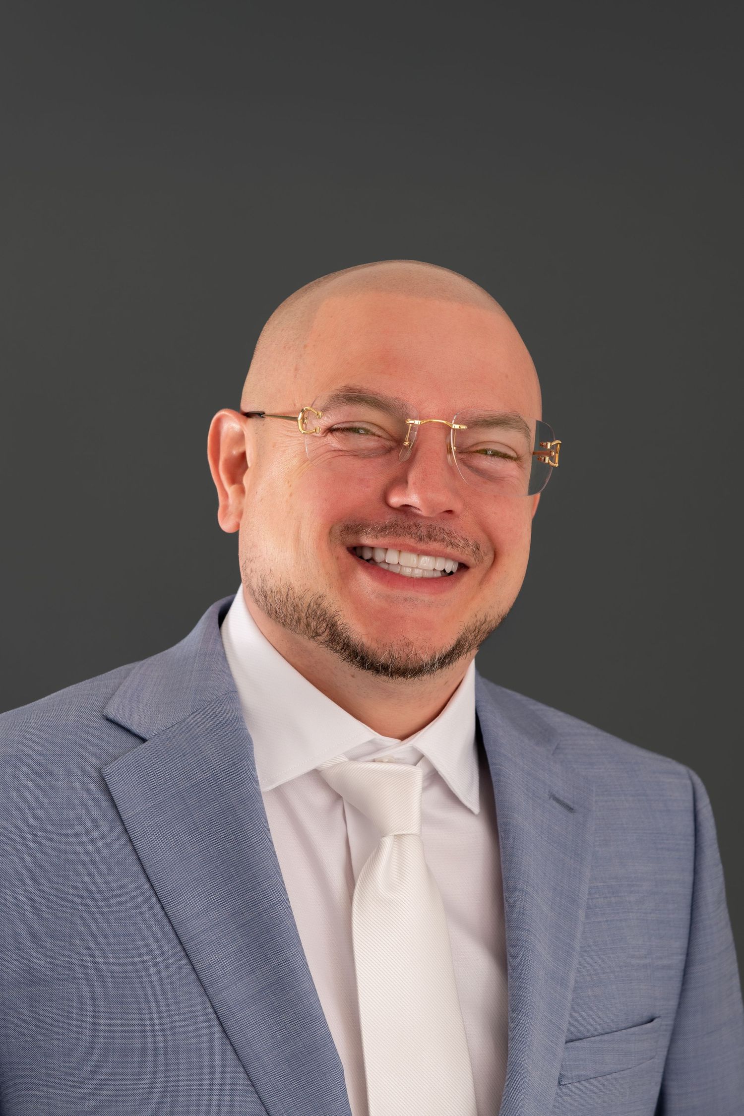 A bald man with glasses poses on a grey backdrop with a grey suit and white shirt, smiling.