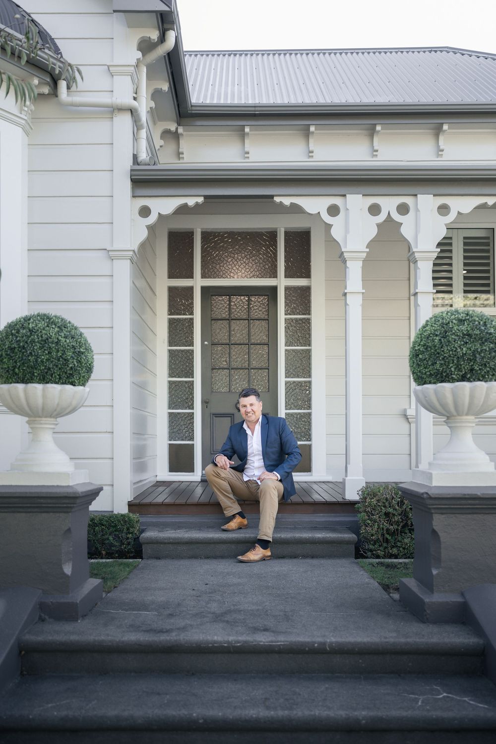 Man in blazer sitting on steps of elegant white Victorian home with ornate veranda and topiary urns.