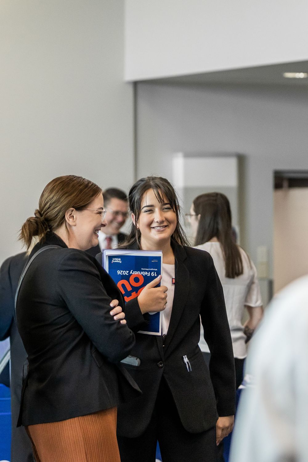 Two professional women in black suits smiling and sharing a document at a business networking event.