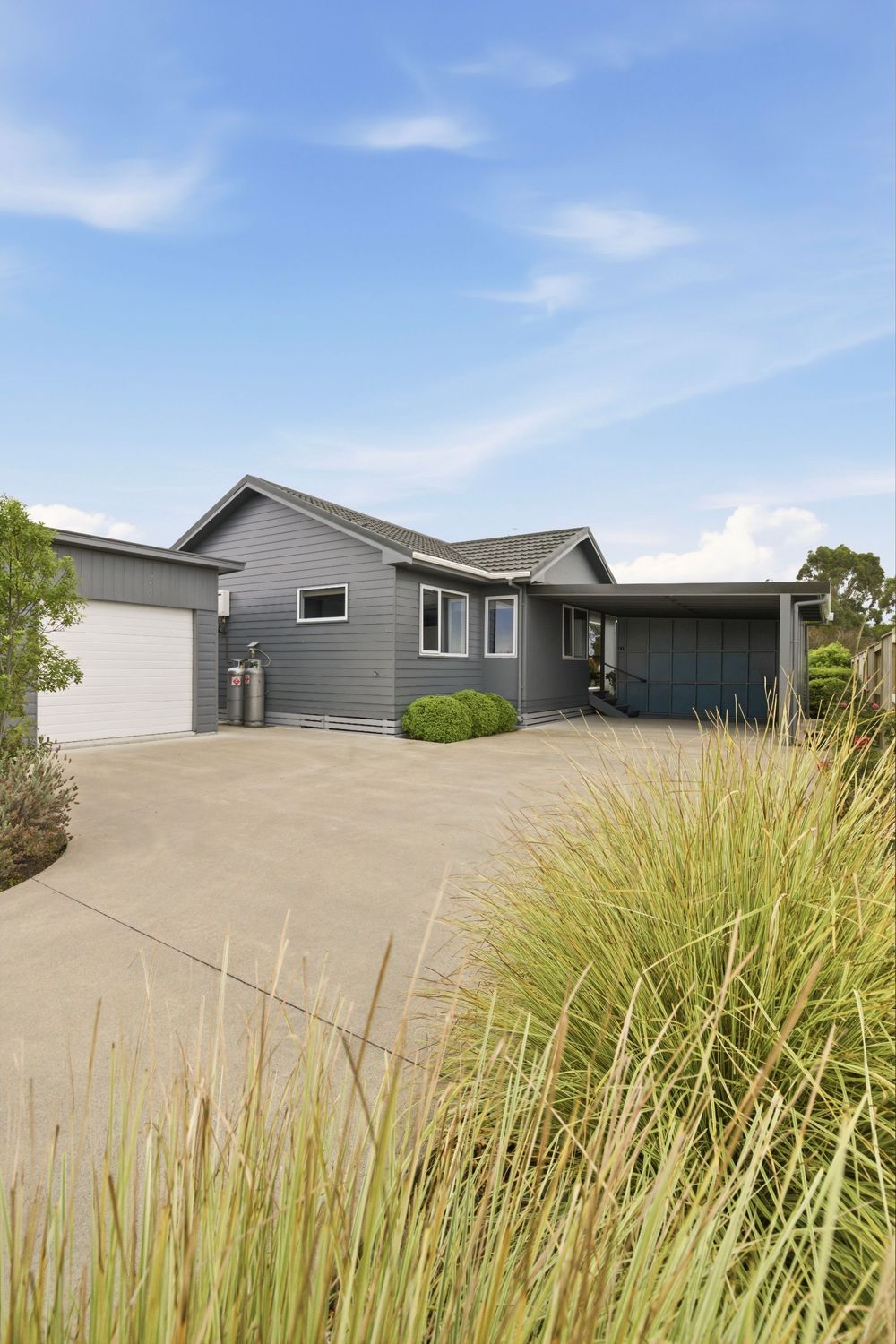Modern gray single-story home with wide concrete driveway, double garage, and ornamental grasses under a blue sky.