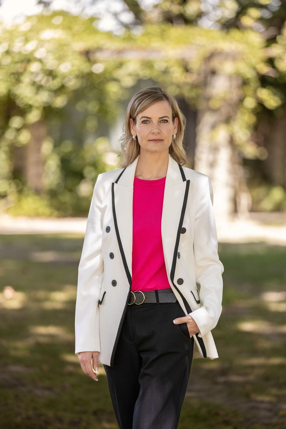Professional woman in white blazer and pink top walking outdoors in a garden setting.