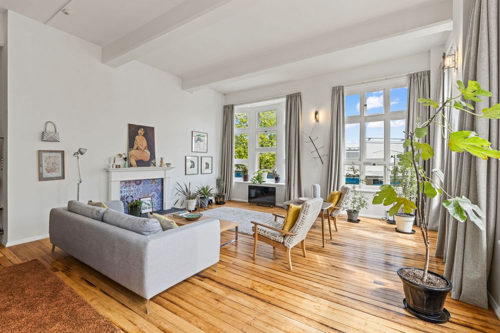 Bright living room with hardwood floors, gray sofa, fireplace, large windows, and lush indoor plants in a modern apartment.