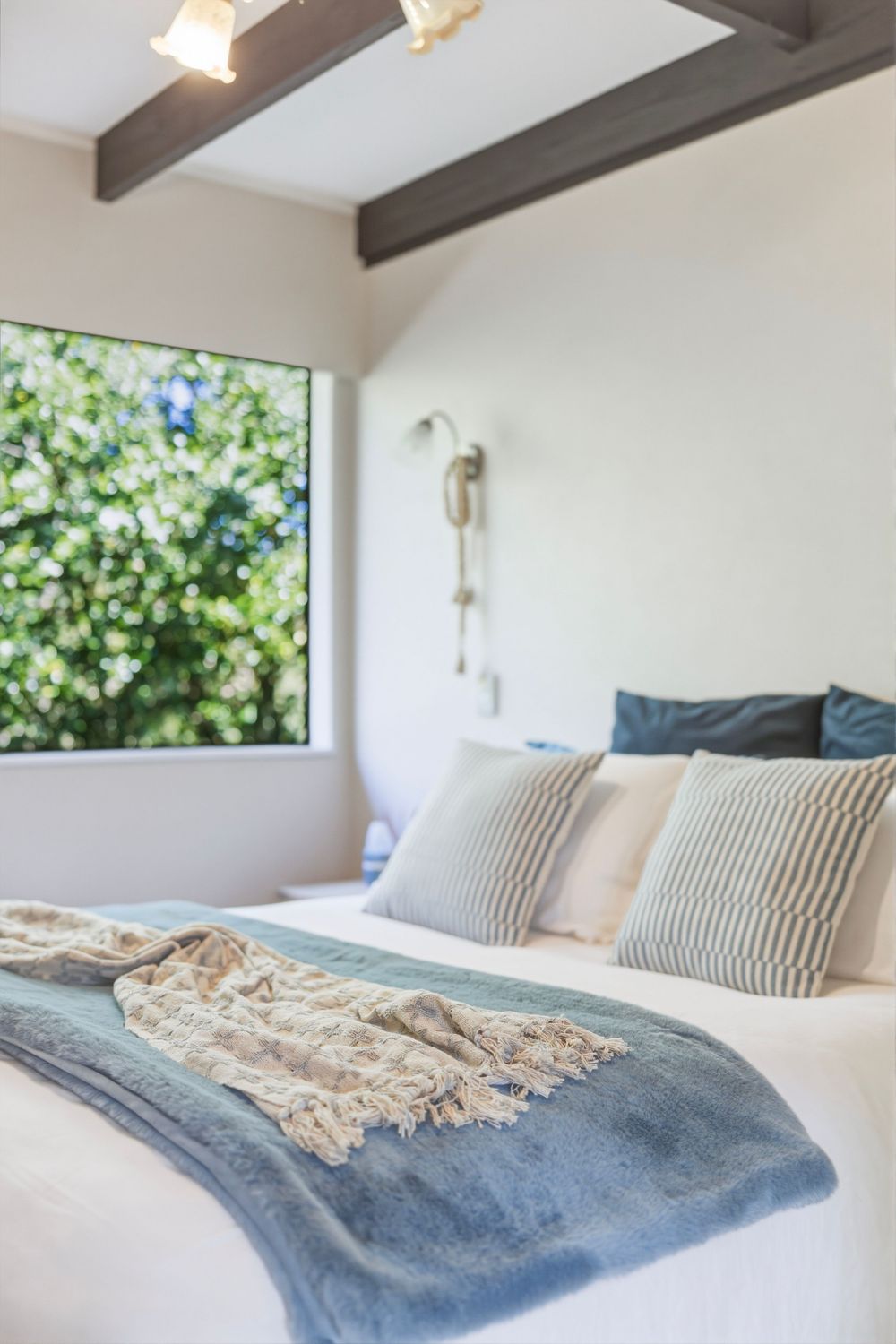 Cozy bedroom with white bedding, blue throw blanket, striped pillows, wooden ceiling beams, and a window with green tree views.
