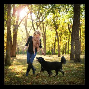 Woman in black top and pink scarf playing with a black dog in a sunlit autumn forest park.