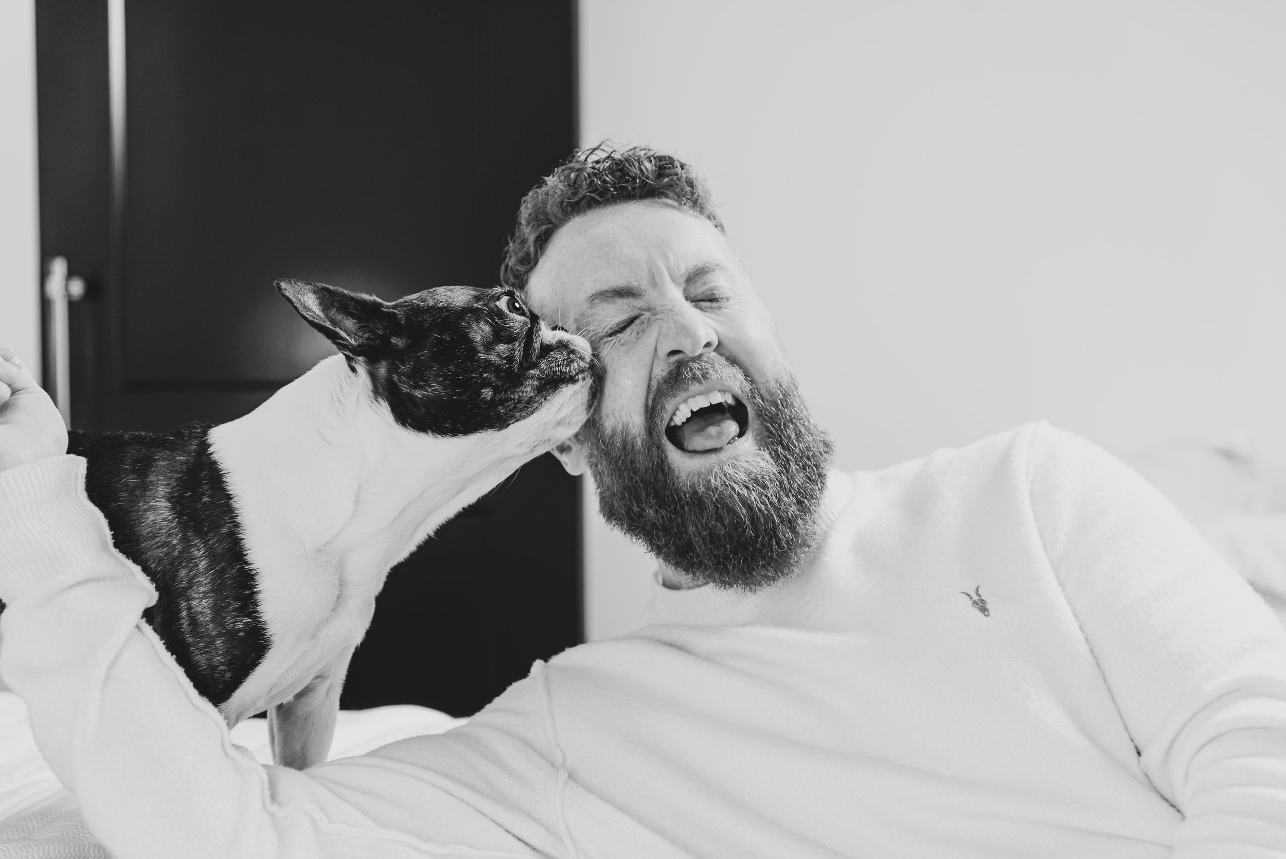 Black and white photo of a bearded man laughing joyfully while a small black cat nuzzles against his face.