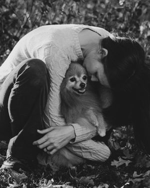 Black and white photo of a person hugging a small Pomeranian dog outdoors among fallen leaves.