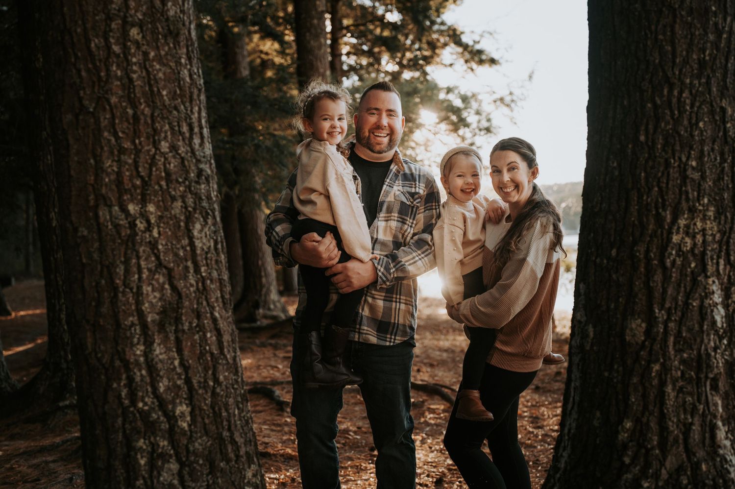Happy family of four posing among tall trees in a sunlit forest, parents holding young children outdoors.