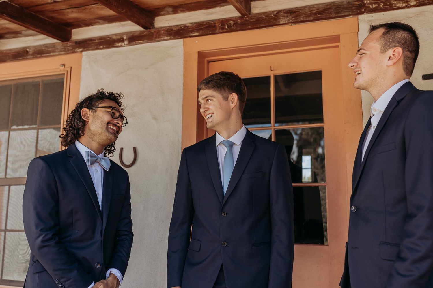 Three men in navy suits smiling and conversing outdoors near a rustic building entrance at Rancho Buena Vista Adobe.