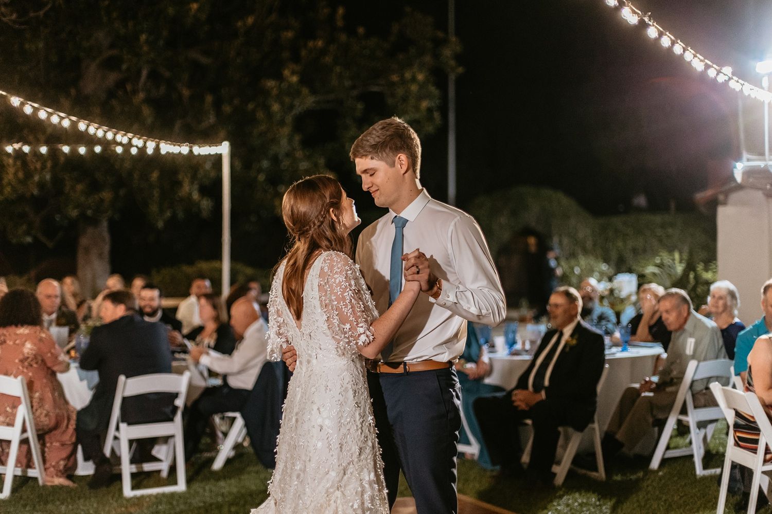Couple sharing first dance at outdoor wedding reception with string lights and seated guests in background at Rancho Buena Vista Adobe.