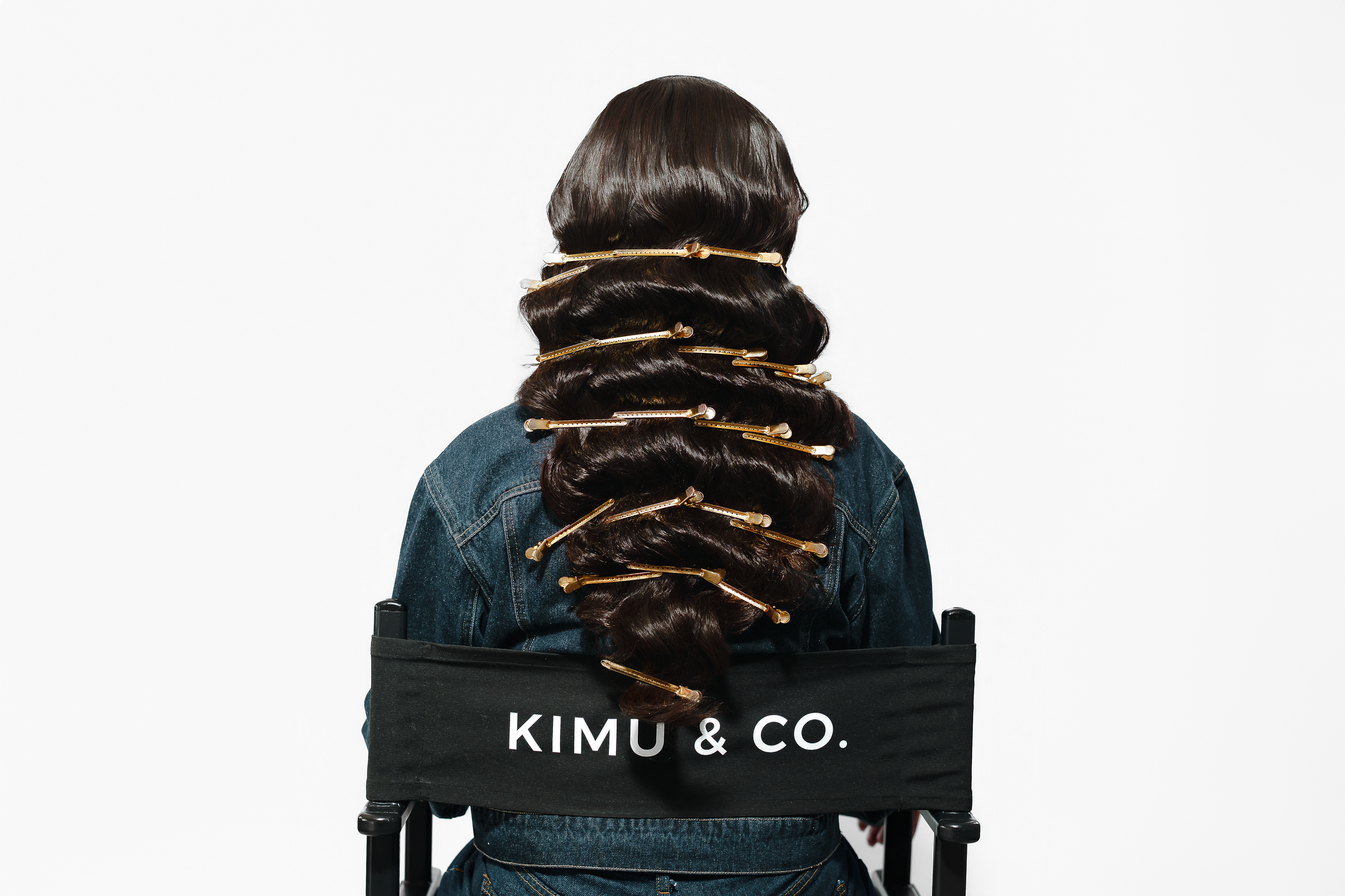 Person seated in director's chair with Kimu & Co. branding, wearing denim jacket with decorative hair accessories.