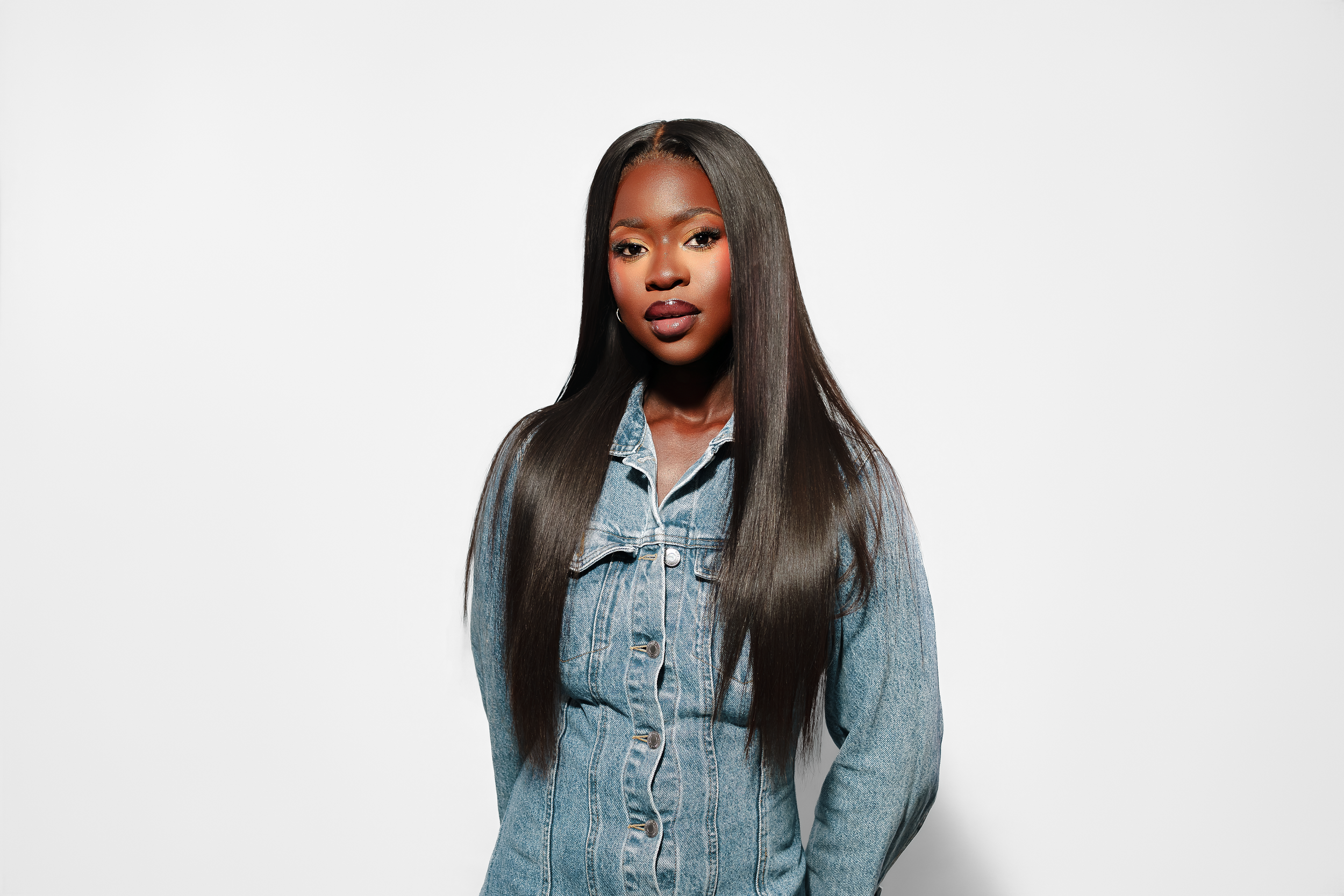 Young Black woman with long straight hair wearing a denim shirt, posing confidently against a white background.