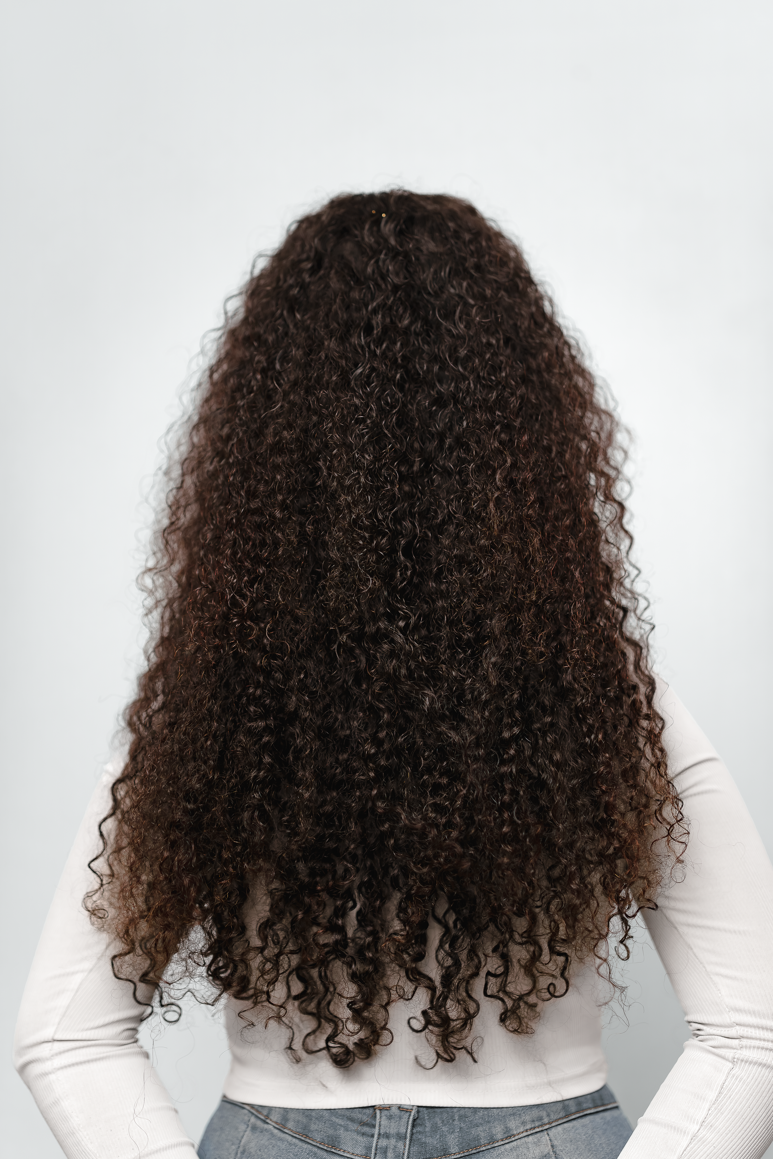 Back view of a woman with long, voluminous dark brown curly hair wearing a white top and jeans.