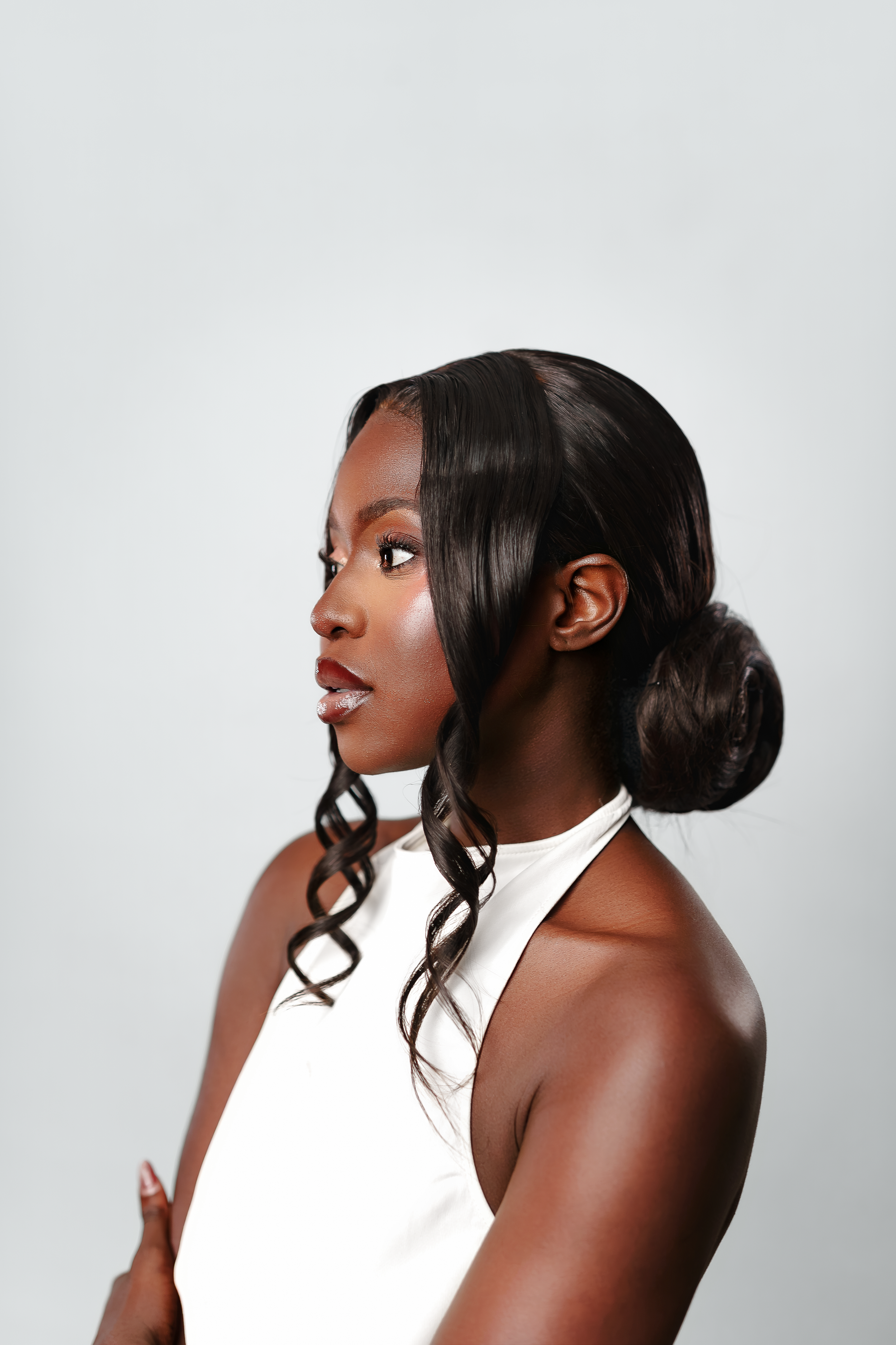 Elegant Black woman in white halter dress with sleek bun hairstyle, posing against light gray background.