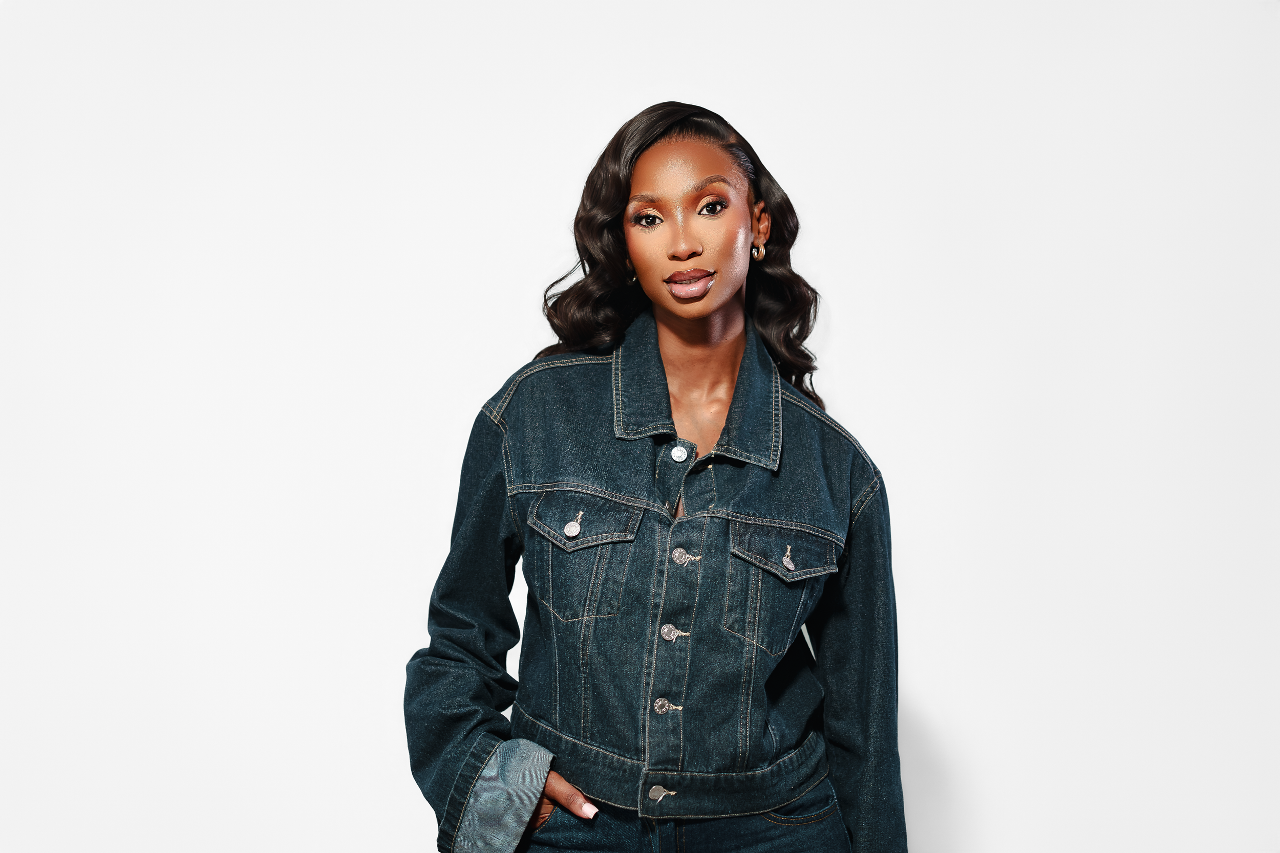 A stylish woman in a dark denim jacket and jeans poses confidently against a white background.