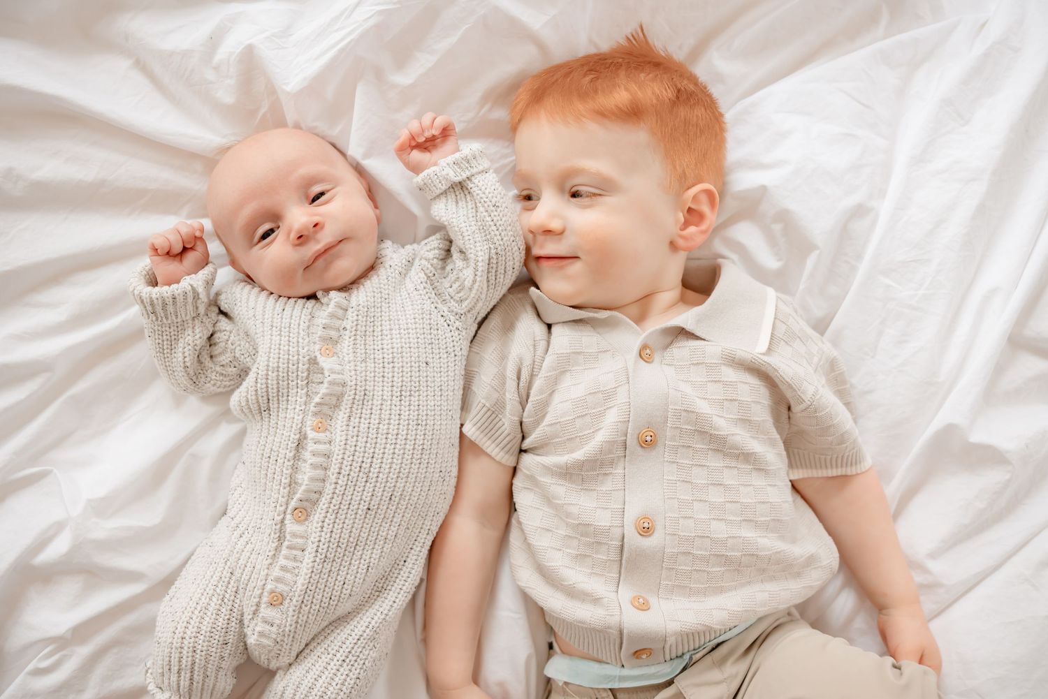 Two babies in matching gray and white outfits lying together on white bedding while playing and laughing.