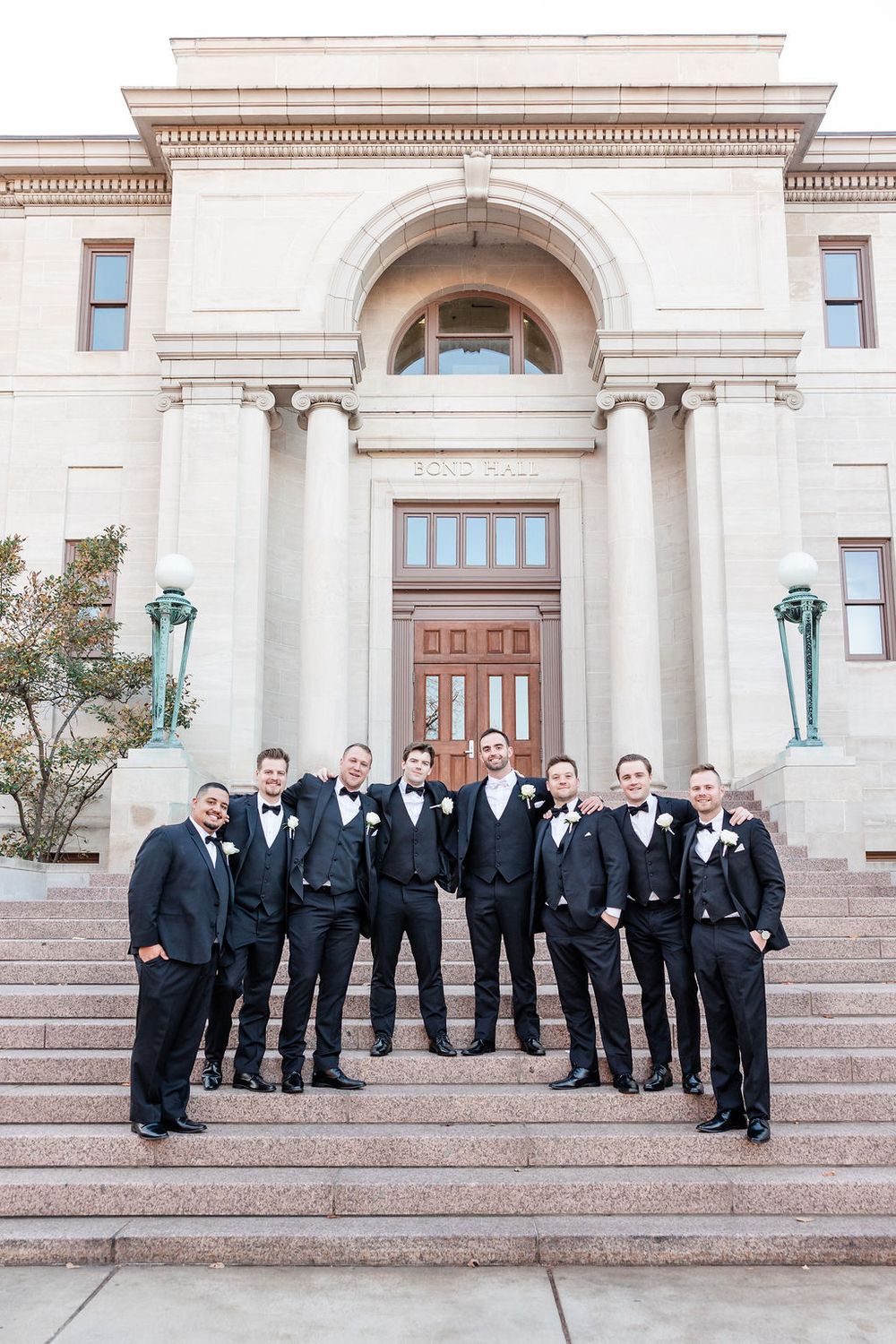 Groomsmen in black tuxedos pose on steps of elegant stone building with columns for wedding photo.