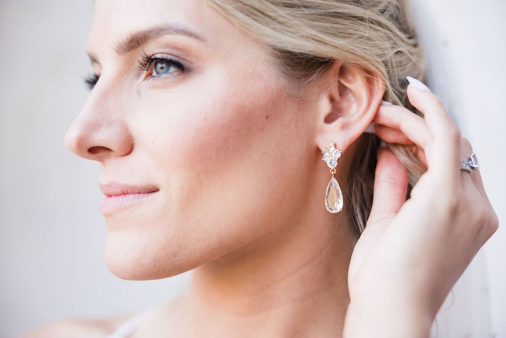Bride adjusting elegant teardrop crystal earring, wearing bridal jewelry with soft makeup and updo hairstyle.