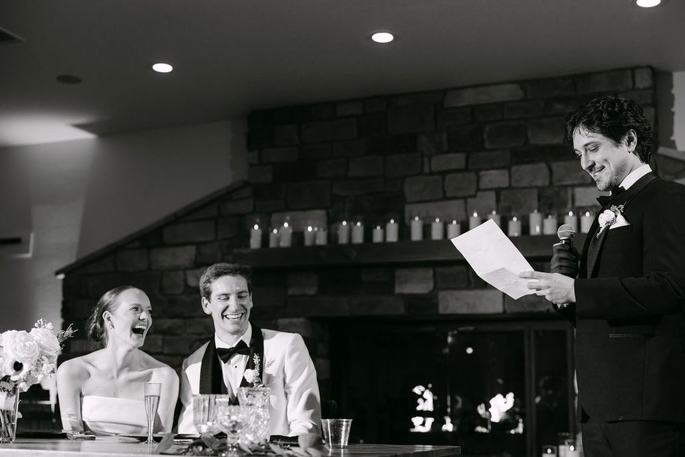 Black and white photo of bride and groom laughing during wedding speech at elegant reception.