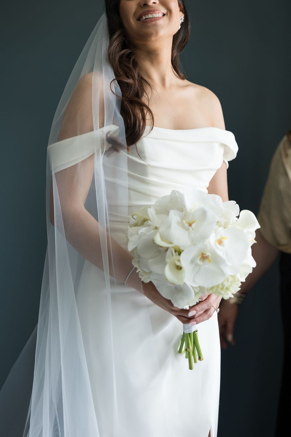 Smiling bride in off-shoulder white gown and veil holding elegant white orchid bouquet against dark background.