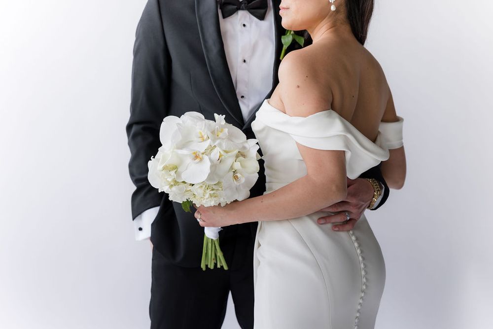 Bride and groom embrace holding white orchid bouquet against white background in classic wedding portrait.