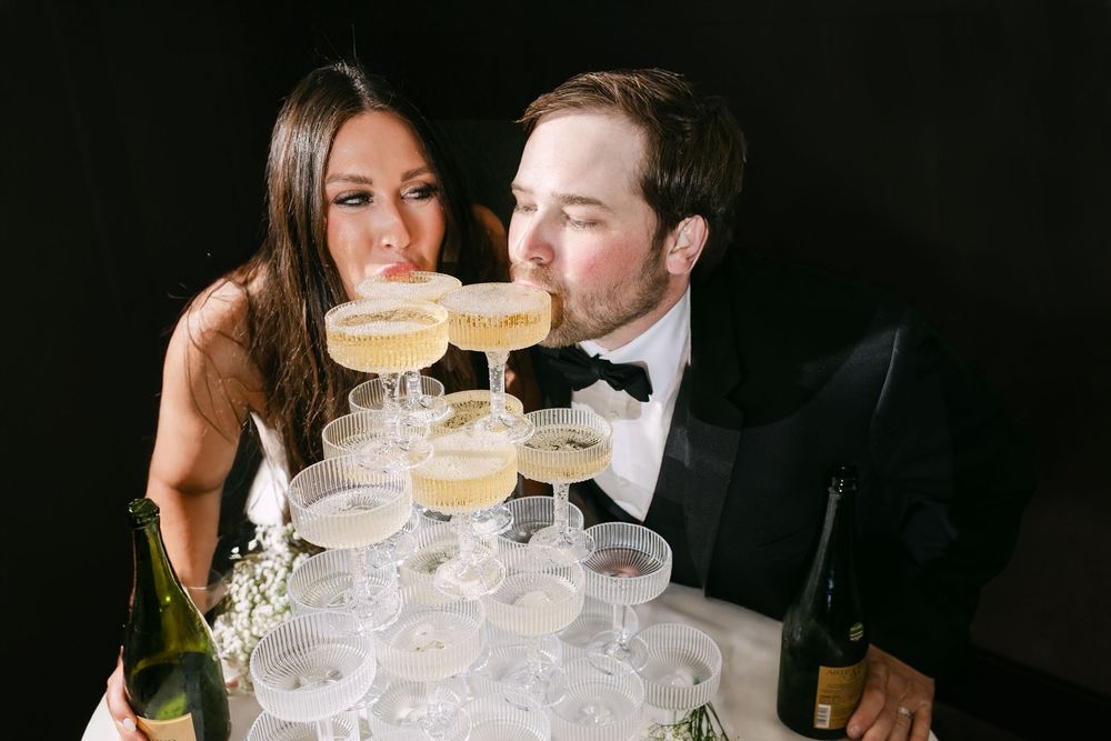 Bride and groom sipping champagne together from a tower of coupe glasses at their wedding reception.