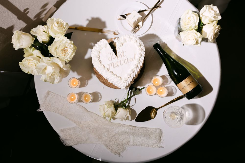 White heart-shaped wedding cake with roses, champagne bottle, candles, and lace on a round white table.