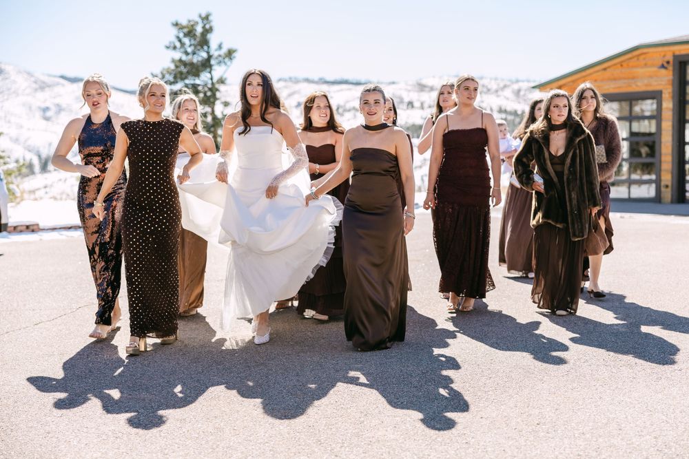 Bride in white gown walks with bridesmaids in dark dresses outdoors at a snowy mountain wedding venue.