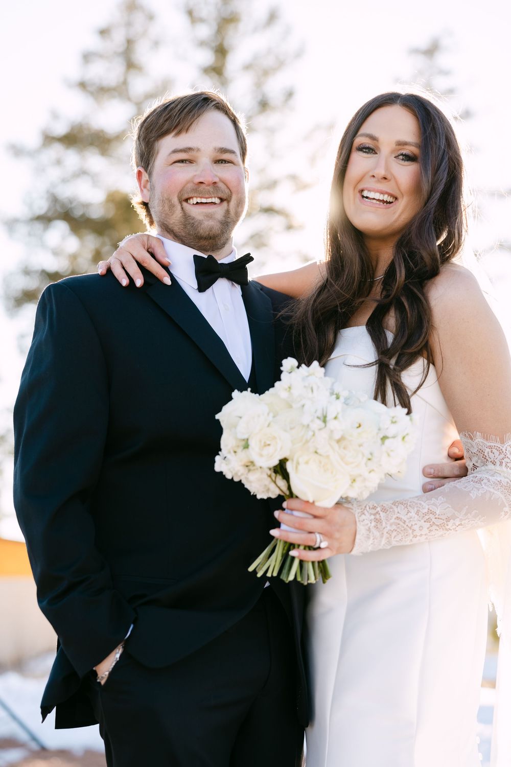 Smiling bride and groom in wedding attire, bride holding white floral bouquet, posing outdoors in elegant formal wear.