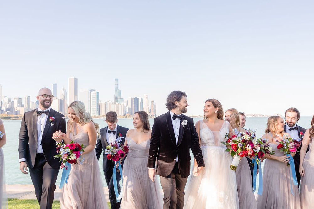 A wedding party walks together along a lakefront with a stunning city skyline backdrop on a bright sunny day.