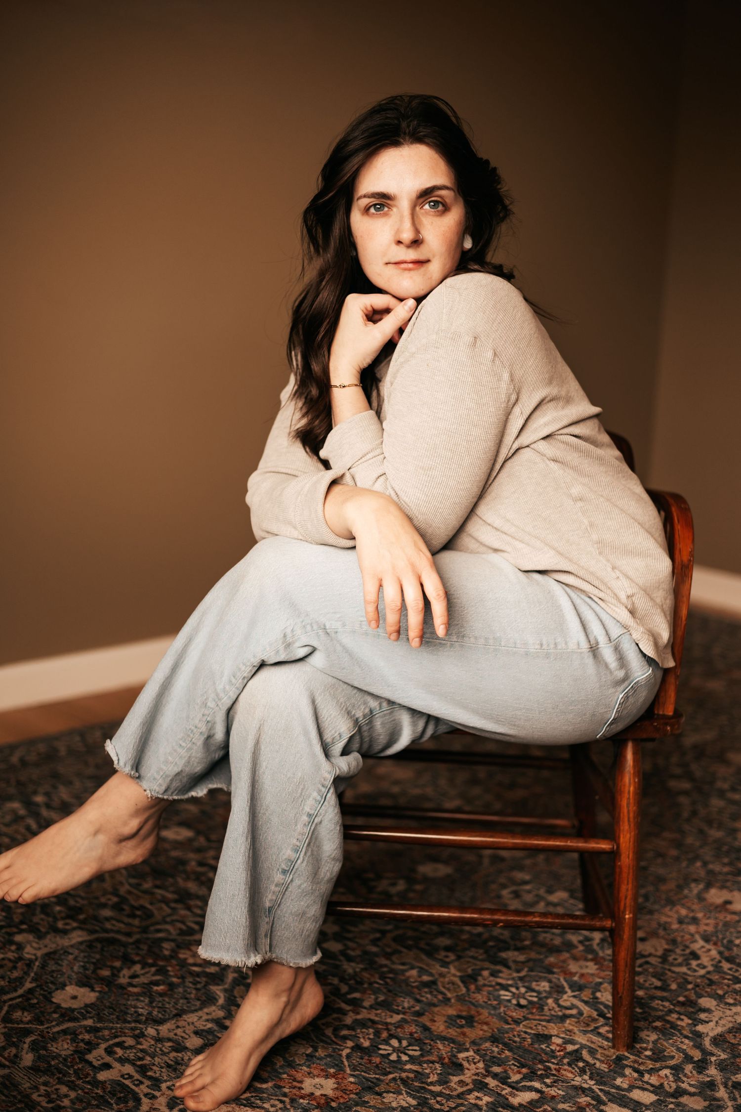 Young woman in beige sweater and light jeans sits barefoot on wooden chair against brown wall |My Postpartum Depression Story