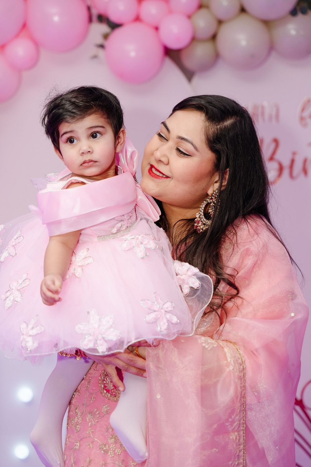 A smiling mother in a pink saree holds her baby girl in a pink floral dress at a pink balloon party backdrop.
