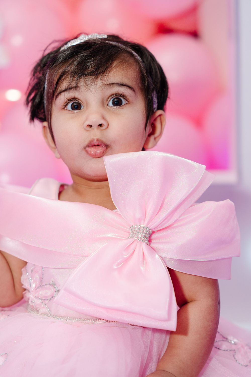 A cute baby girl in a pink bow dress with a tiara makes a funny face against a pink balloon background.
