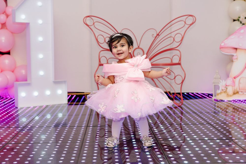 A toddler girl in a pink dress with butterfly wings dances on a sparkly dance floor at a fairy birthday party.