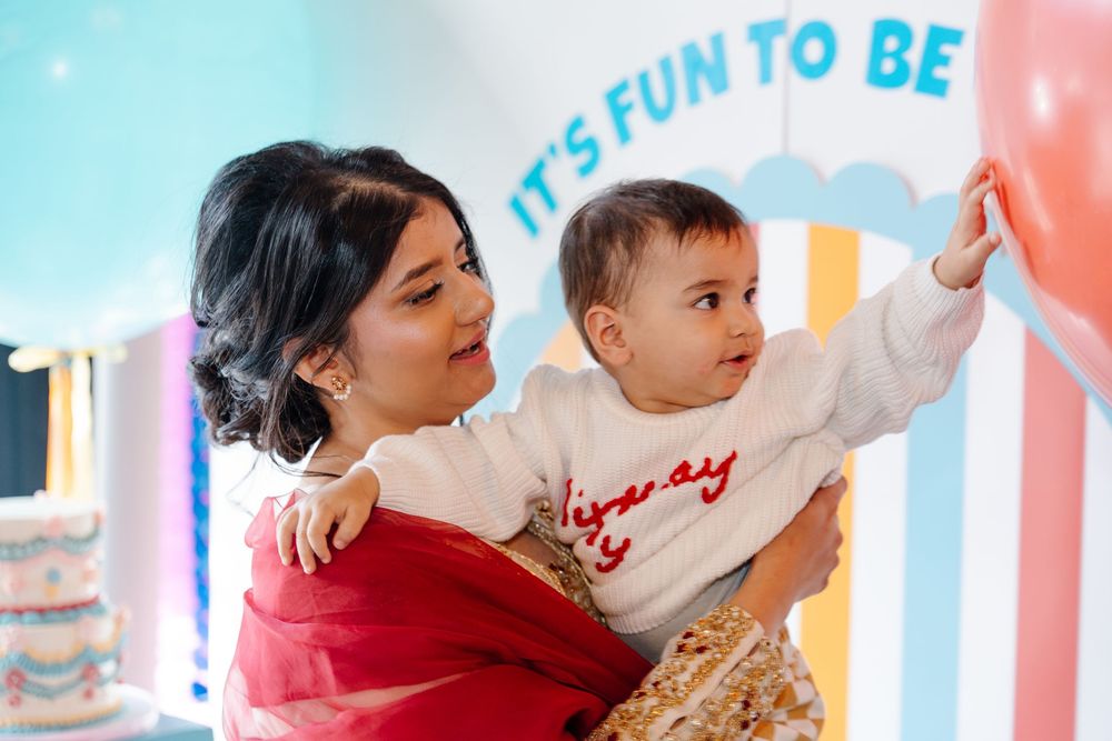 Mother in red dress holds baby reaching for balloon at colorful first birthday party.