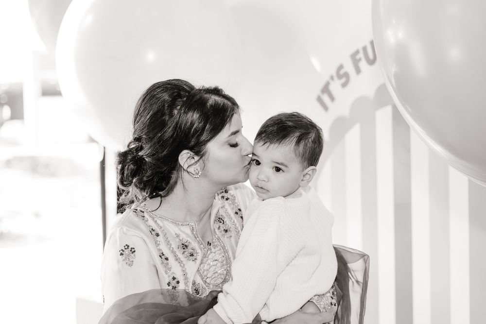 Black and white photo of mother kissing baby's cheek at first birthday balloon celebration.