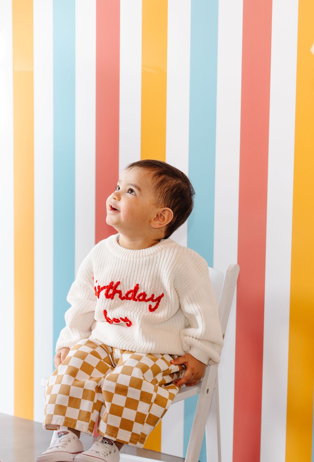 A baby boy in a white 'Birthday Boy' sweater and checkered pants sits on a white chair against a colorful striped backdrop.
