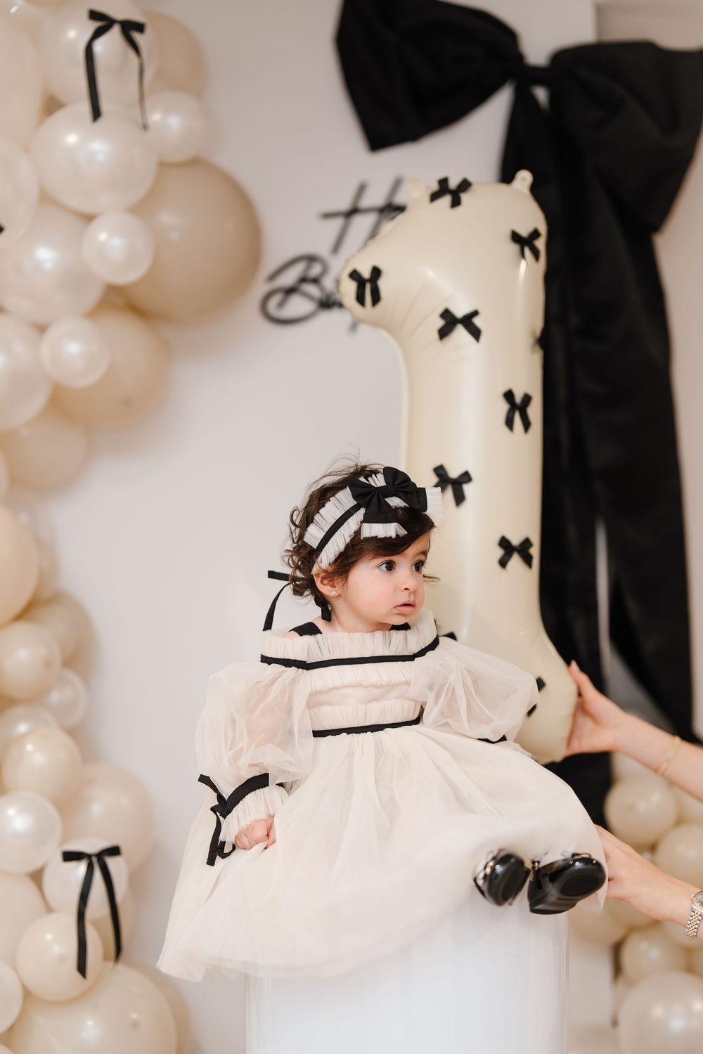 A baby girl in a white and black bow dress sits beside a number one balloon at her first birthday party.