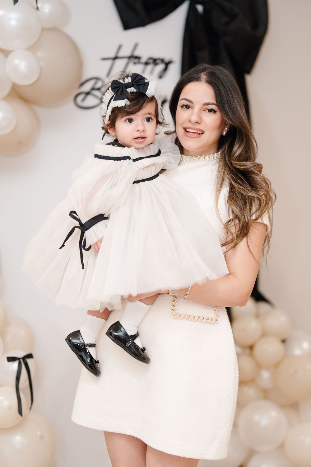 A mother in a white dress holds her baby girl in a matching outfit at a black and white birthday celebration.