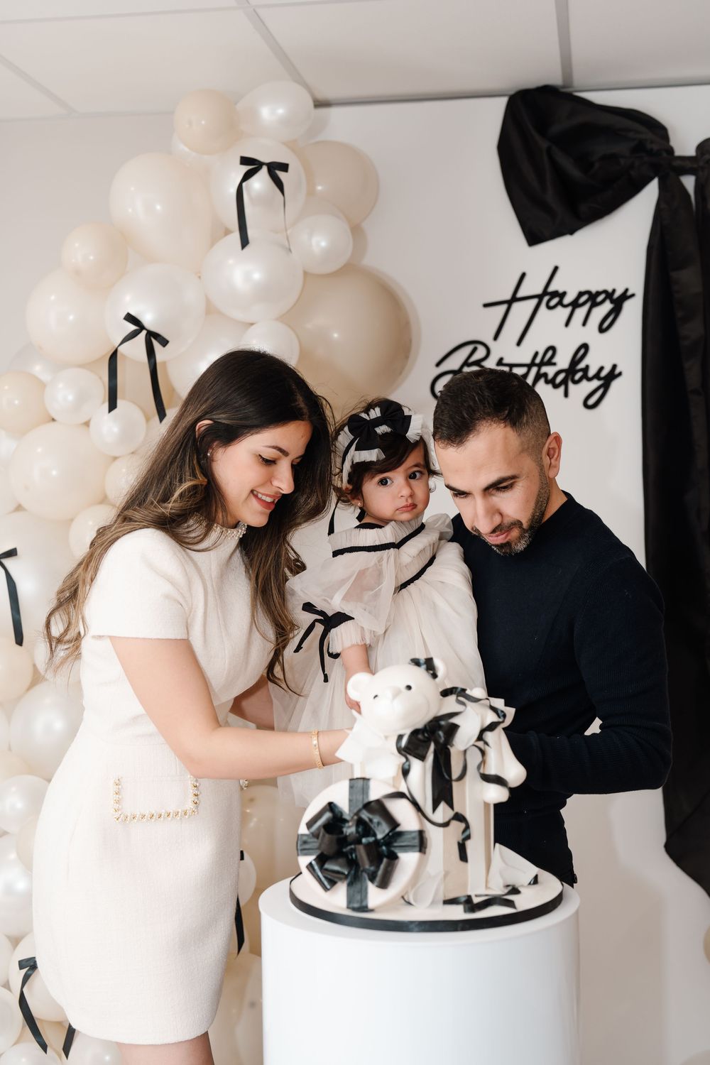 A family admires a black and white teddy bear birthday cake at an elegant monochrome first birthday party.
