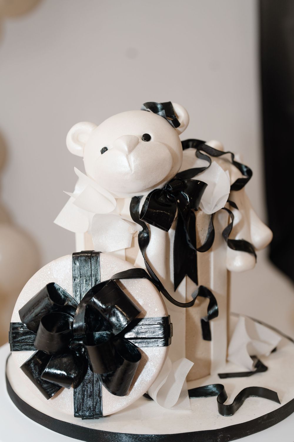 An elegant black and white first birthday cake featuring a teddy bear topper and decorative ribbon bows.